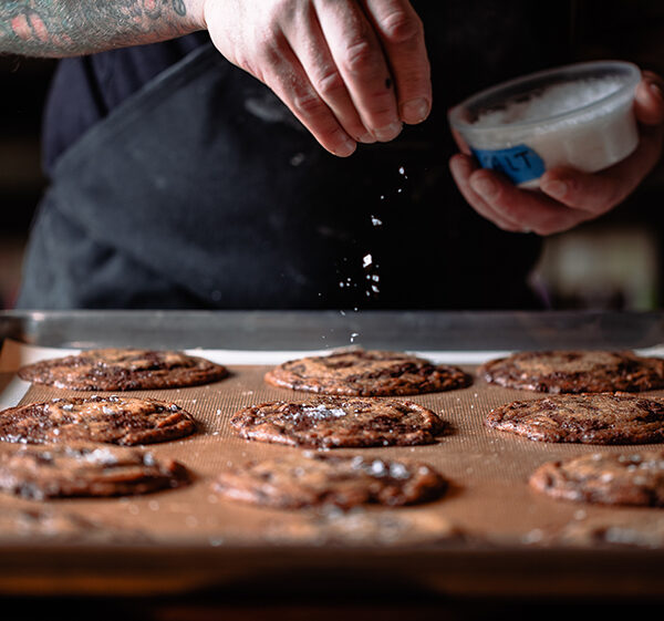 Cookies being finished with flaky salt on a baking sheet